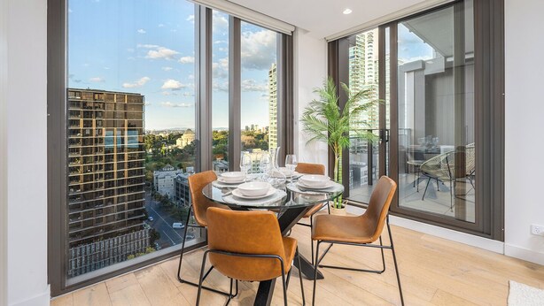 A dining area with a table set for four, large windows, and a view of a cityscape. - Luxury Sky High Apartment with Pool Gym & Parking (South Melbourne)