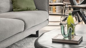 A modern living room with a grey sofa, a green pillow, a glass table with a bottle and glasses, and a view of buildings through large windows. - Luxury Sky High Apartment with Pool Gym & Parking (South Melbourne)