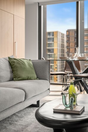Ett modernt vardagsrum med en grå soffa, en grön kudde, ett glasbord med en flaska och glas, och en utsikt över byggnader genom stora fönster.