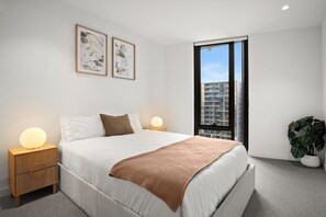 Ein modernes Schlafzimmer mit einem großen Bett, Nachttischlampen, Blick auf Gebäude durch ein Fenster und einer Zimmerpflanze.