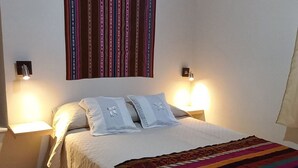 Une chambre à coucher avec un lit, deux lampes de chevet, une couverture rayée aux couleurs vives et un luminaire suspendu aux motifs géométriques.