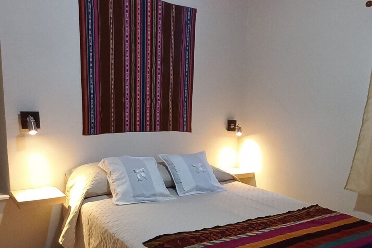 Un dormitorio con cama, dos lámparas de noche, una manta a rayas coloridas y una lámpara colgante de diseño geométrico.
