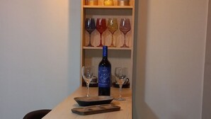 Un coin bar avec un comptoir en bois, deux tabourets, une bouteille de vin et des étagères où sont disposés des verres et des plantes.