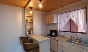Cocina compacta con barra, armarios de madera, cafetera y ventana con cortina.
