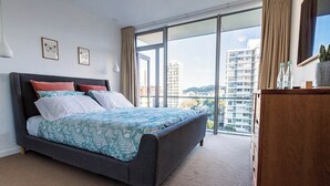 A modern bedroom with a large bed, a wooden dresser, and a city view through floor-to-ceiling windows. - Executive Apartment - Great Location Close to Attractions, Shops & Eateries (Wellington)
