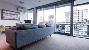 A modern living room with a grey sofa, a framed picture on the wall, and a large window offering a city view. - Executive Apartment - Great Location Close to Attractions, Shops & Eateries (Wellington)