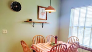 A dining area with a checkered tablecloth, wooden chairs, a wall clock, a framed picture, and a hanging light fixture.