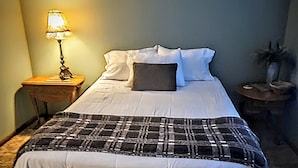 A neatly made bed with a plaid blanket, a small bedside table with a lamp, and a framed picture on the wall.
