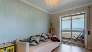 Ein Zimmer mit einer Couch, einer Gitarre, einem Tisch mit einem Gesellschaftsspiel und Blick auf den Strand durch eine Schiebetür.