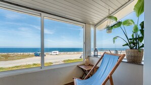 Ein Zimmer mit großem Fenster mit Blick auf einen Strand, ein Holzstuhl, ein Blumentopf und ein kleiner Tisch mit einer Laterne.
