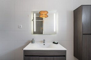 Une salle de bain avec un lavabo blanc, un miroir, un luminaire en osier et un meuble en bois foncé.