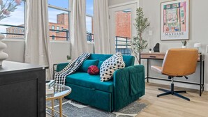 A modern living room with a teal sofa, a round coffee table, and a desk with a chair. A large window offers a view of buildings outside. - Luxury 1BR|1BA in the Heart of Fishtown (Philadelphia)