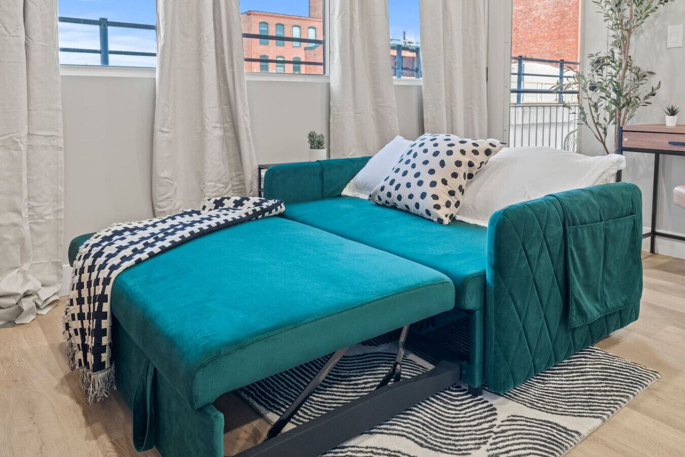 A teal sofa bed with a patterned throw blanket, white pillows, and a black and white striped rug.