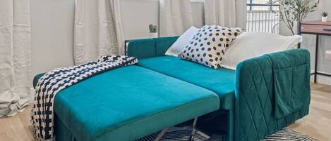 A teal sofa bed with a patterned throw blanket, white pillows, and a black and white striped rug.