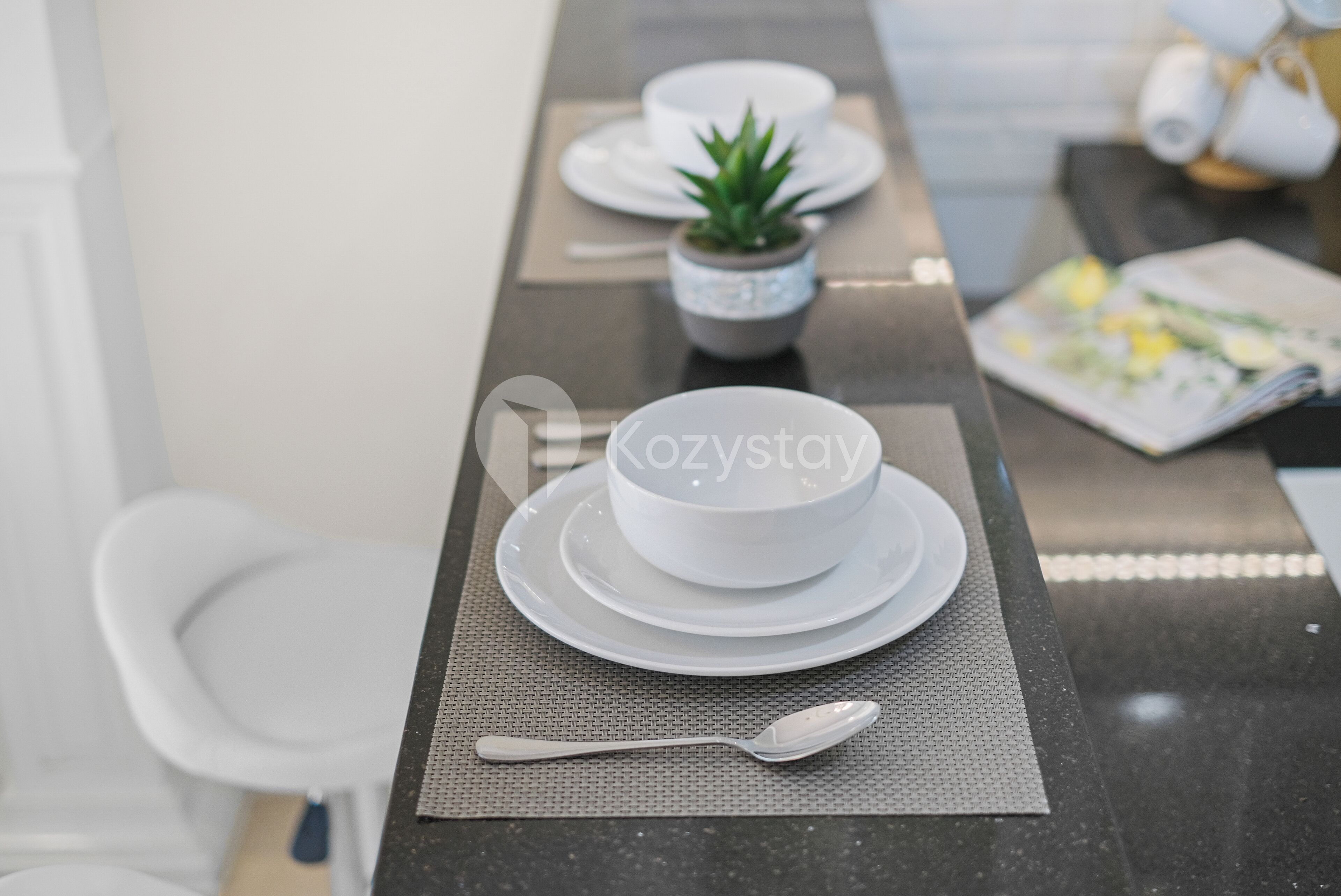 A modern dining area with a white table set for two, a small potted plant, and a magazine on the table.