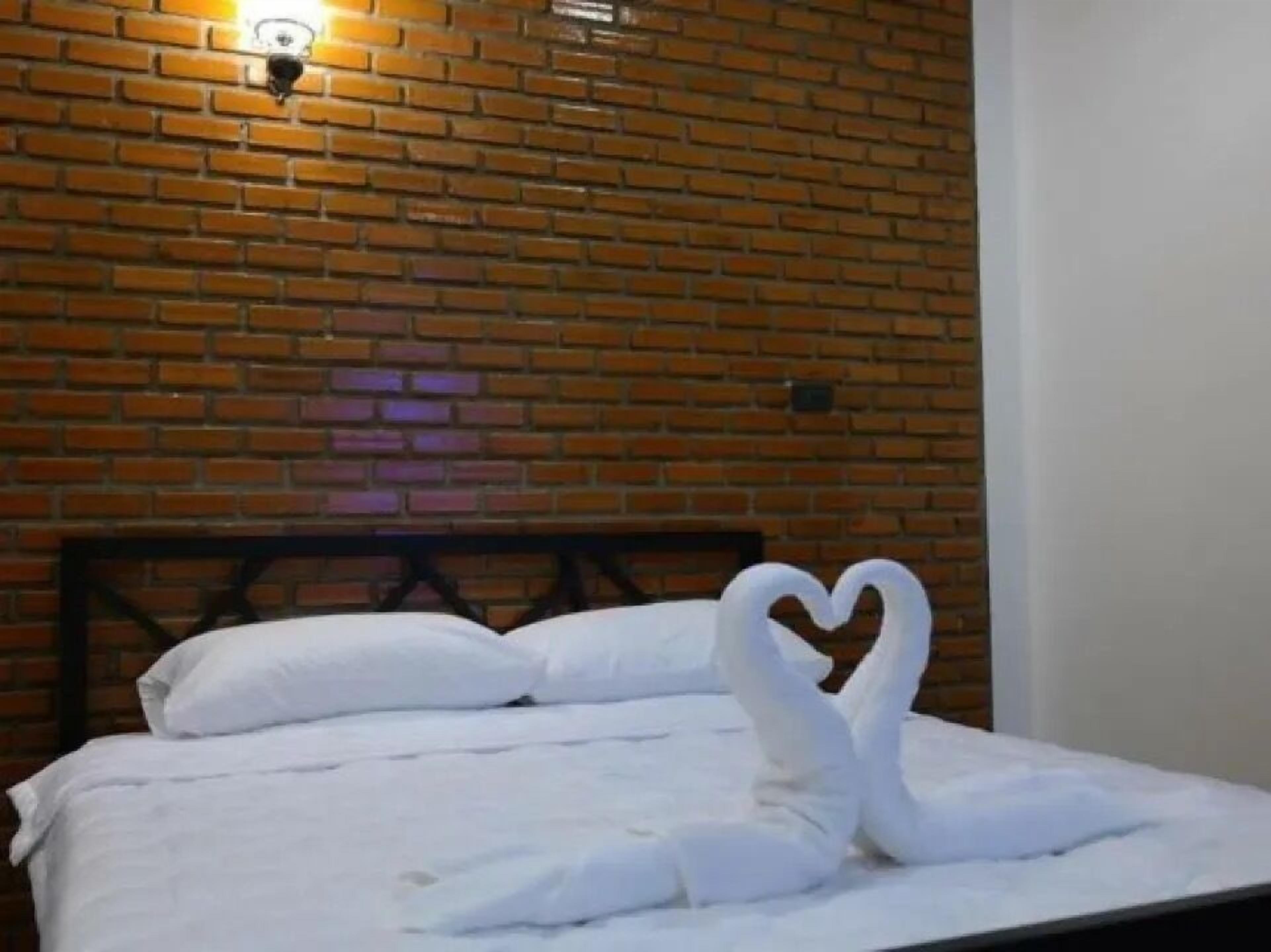 A hotel room with a brick wall, a bed with white bedding, and a heart-shaped towel arrangement.