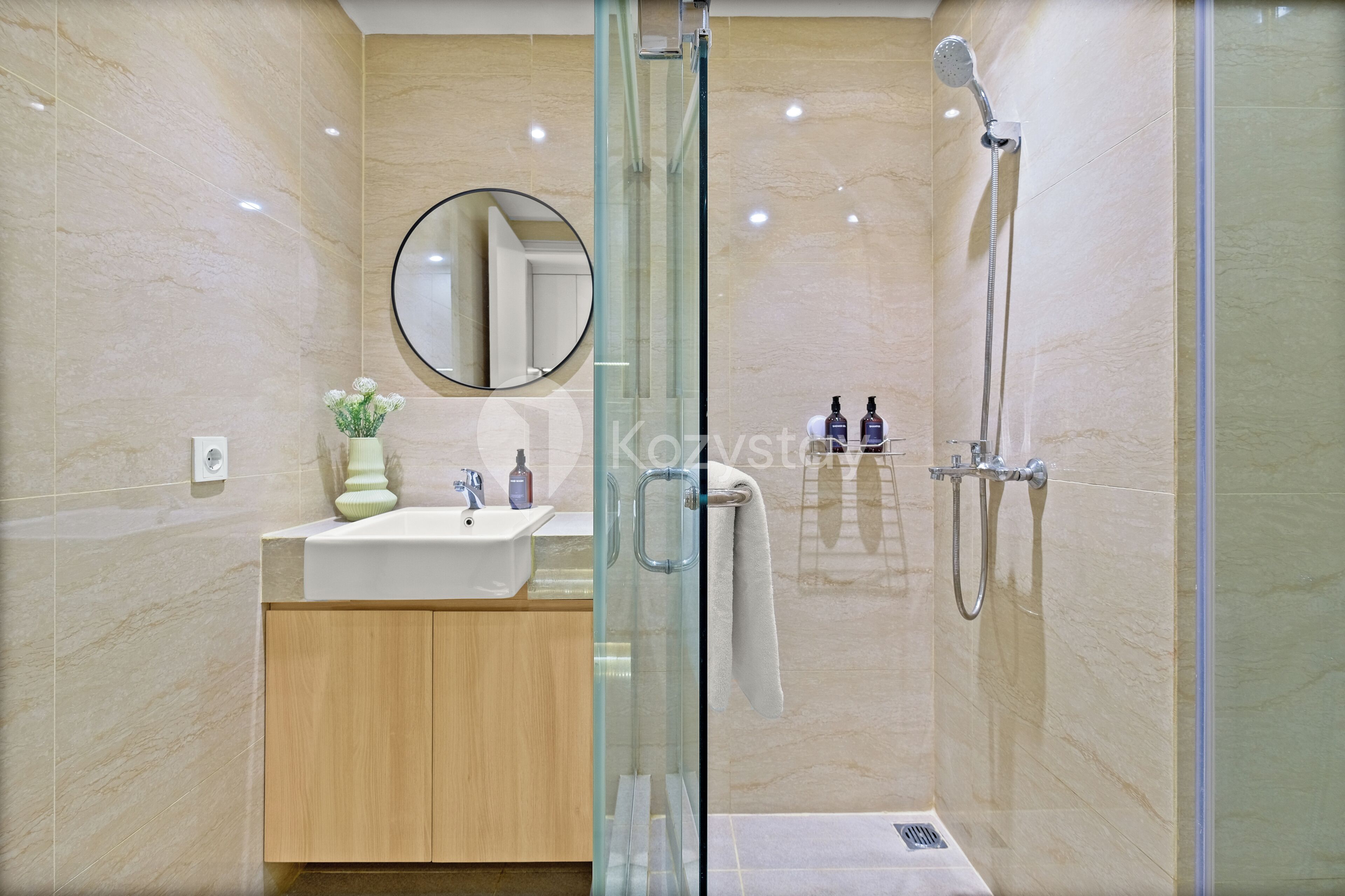 A modern bathroom with a glass-enclosed shower, a round mirror, and a wooden vanity with a sink and two bottles of soap.
