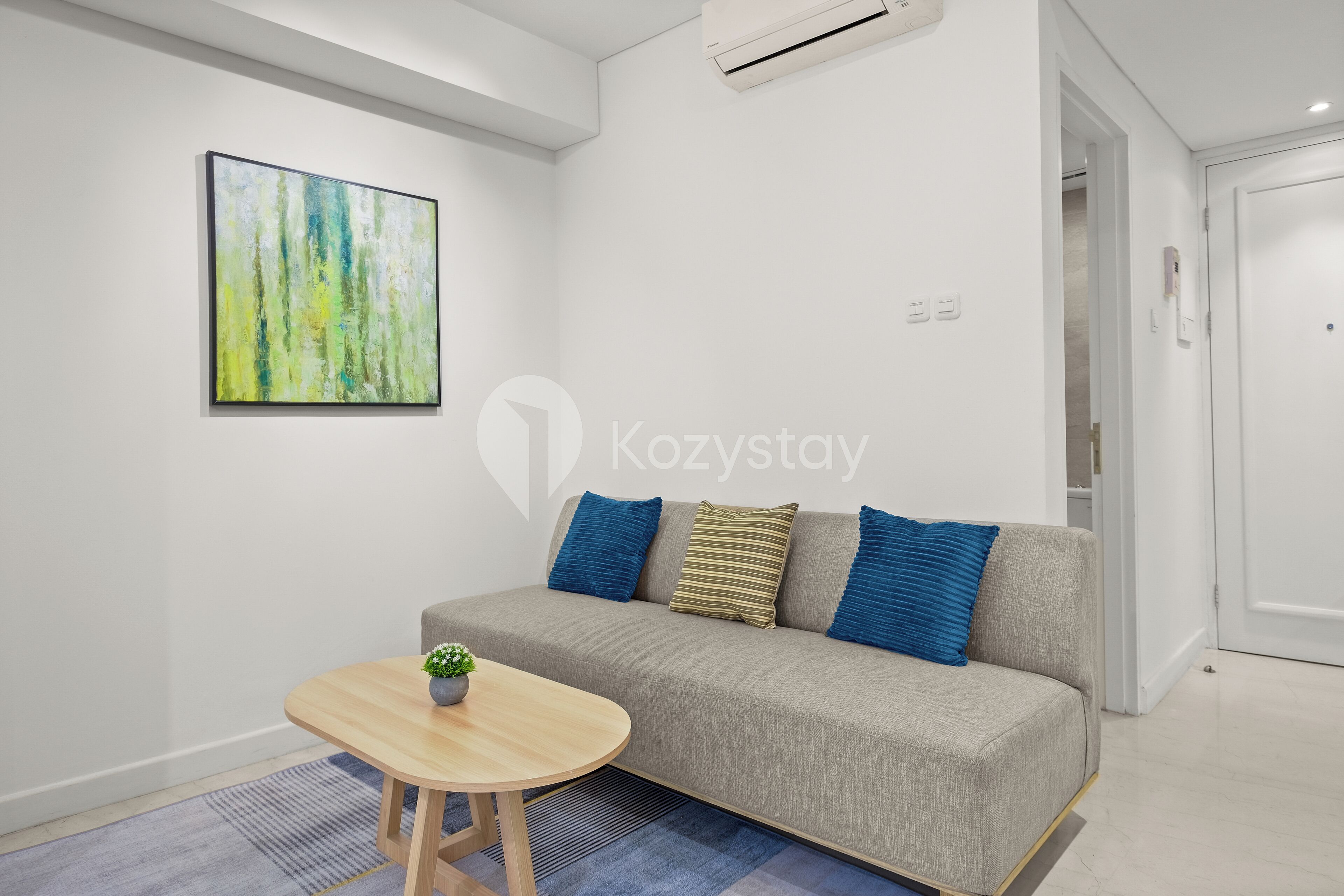 A modern living room with a grey sofa, blue cushions, a small round wooden table with a plant, and a colorful abstract painting on the wall.
