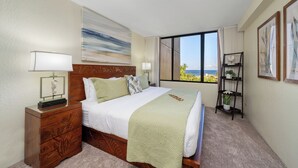 A hotel room with a large bed, a nightstand with a lamp, a view of the ocean through a large window, and a small shelf with plants. - Oceanview 2BD Condo Steps to the Pool - Kaanapali Shores 529 (Lahaina)