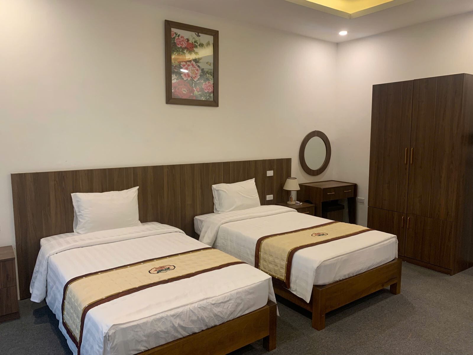 A hotel room with two beds, a wooden headboard, a nightstand with a lamp, a wardrobe, and a painting on the wall.