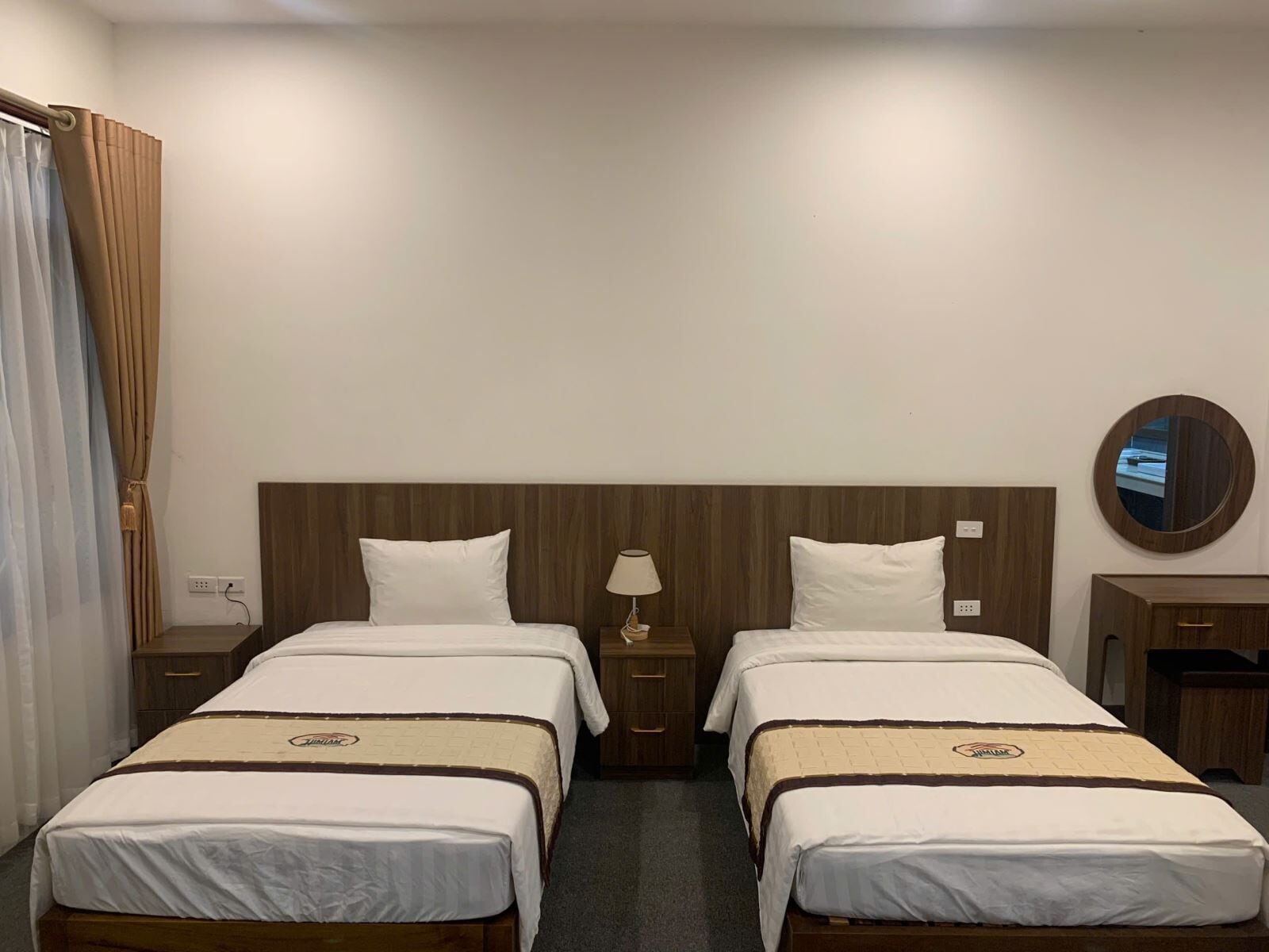 Habitación de hotel con dos camas individuales, cabecera de madera, mesita de noche con lámpara, cómoda pequeña y un espejo redondo en la pared.