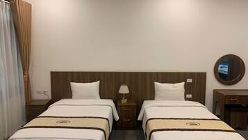 Habitación de hotel con dos camas individuales, cabecera de madera, mesita de noche con lámpara, cómoda pequeña y un espejo redondo en la pared.