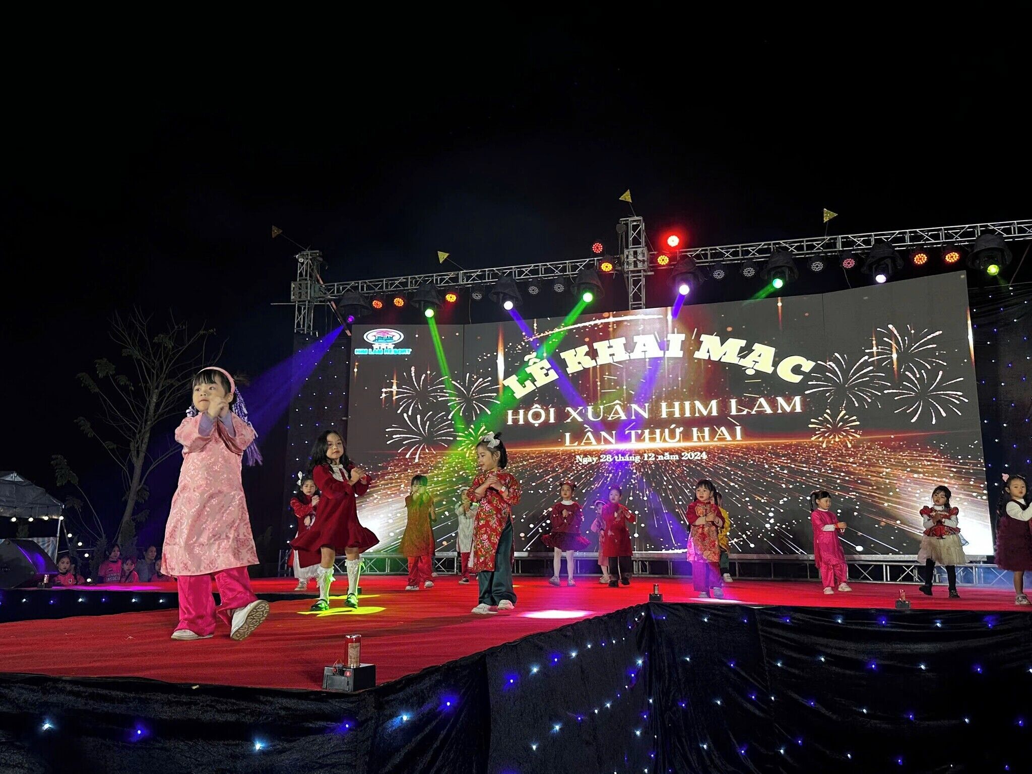 Um palco com artistas em trajes típicos, um grande cenário iluminado e iluminação cênica.