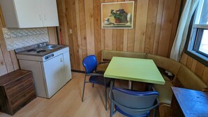 A compact kitchen with a stove, sink, and cabinets, a green table with two chairs, and a wooden wall with a painting.