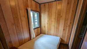 A small, single-bed room with wooden panel walls, a window with curtains, and a small wooden nightstand.