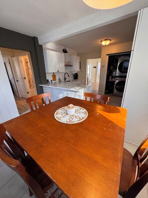 A dining area with a wooden table and chairs, a kitchen in the background, and a laundry room to the right.