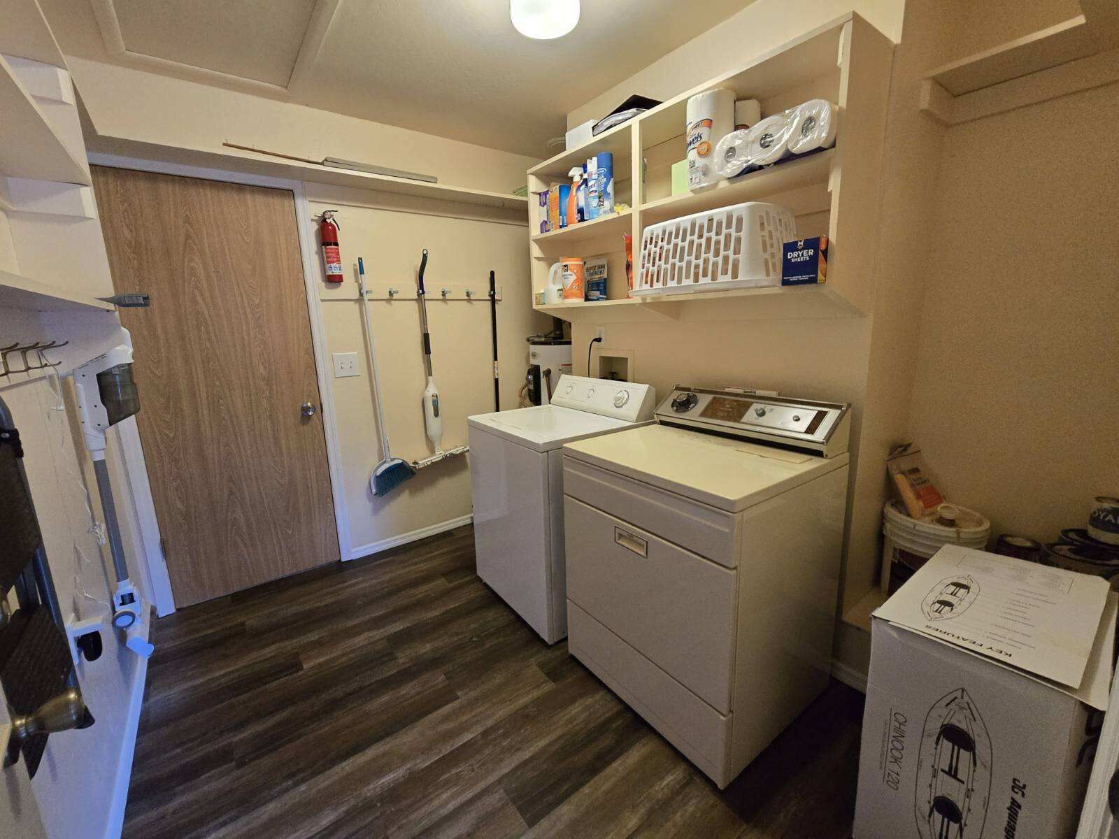 Een wasserette met een wasmachine, droger en planken vol schoonmaakspullen.
