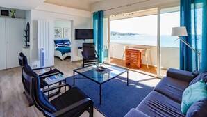 Una sala de estar moderna con un sofá, una mesa de centro y un televisor. Hay un balcón con vista al mar.