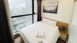 A hotel room with a large window, a bed with white linens, a nightstand with a lamp, and a painting on the wall. - Vinhomes Skylake (Hanoi)
