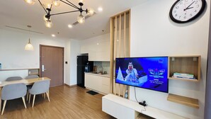 A modern living room with a flat-screen TV, wooden flooring, a dining area with chairs, and a kitchen with a refrigerator. - Vinhomes Skylake (Hanoi)