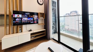 A modern living room with a flat-screen TV displaying various streaming services, a clock on the wall, and a view of a cityscape through large windows. - Vinhomes Skylake (Hanoi)