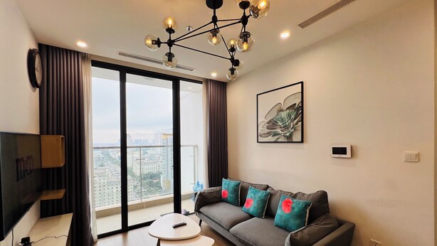 A modern living room with a grey sofa, a coffee table, and a city view through large windows. - Vinhomes Skylake (Hanoi)