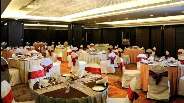 Une salle de banquet avec des tables rondes dressées pour un événement formel, agrémentée de rubans blancs et rouges sur les chaises et surmontée d’un grand lustre.