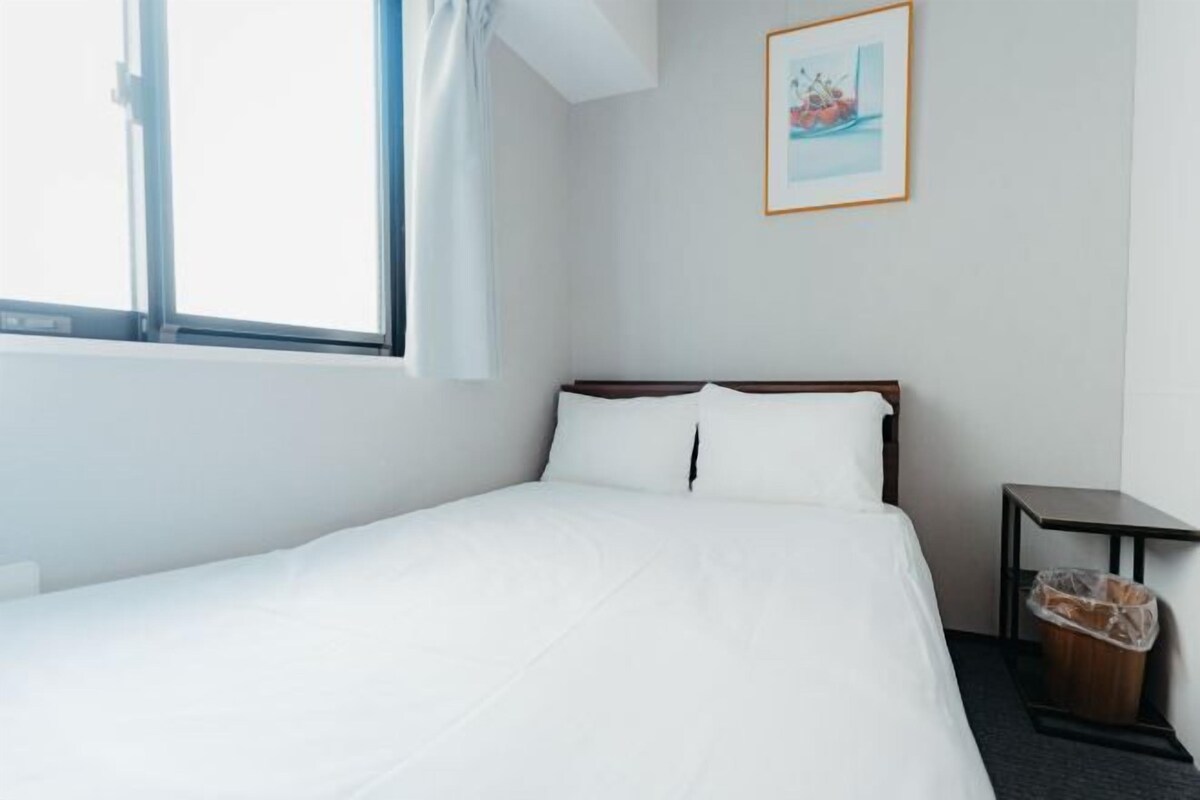 Una habitación de hotel con cama, ventana, un cuadro en la pared y un cubo de basura.