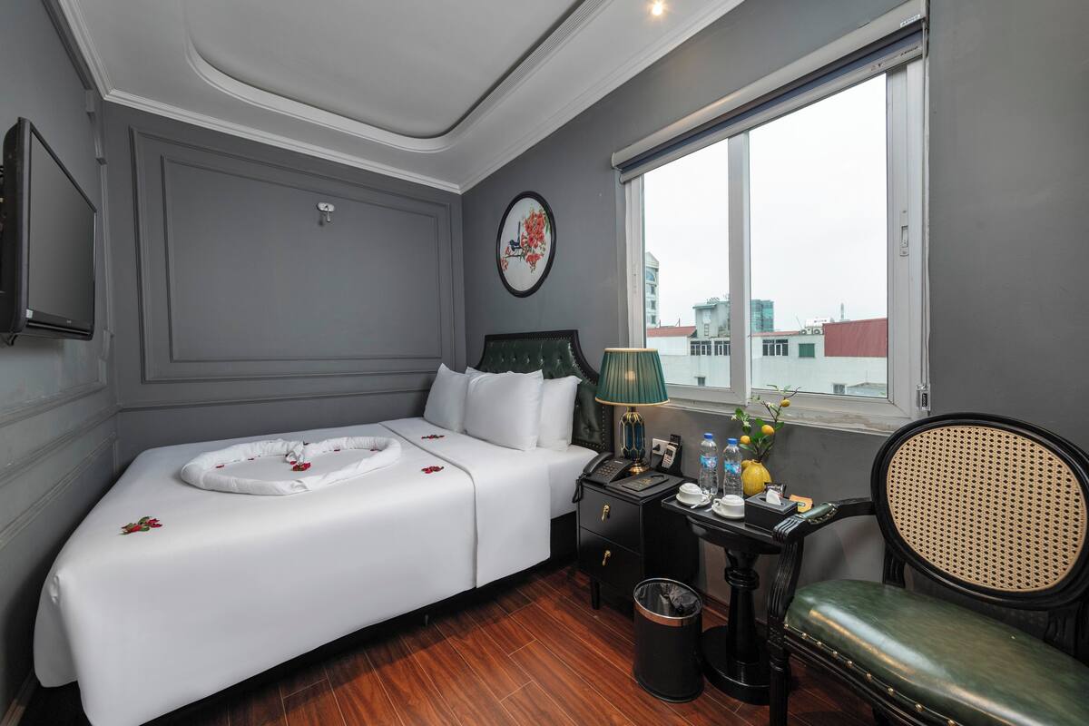 A hotel room with a bed, a desk with a lamp, a chair, a trash bin, and a window with a view of buildings.