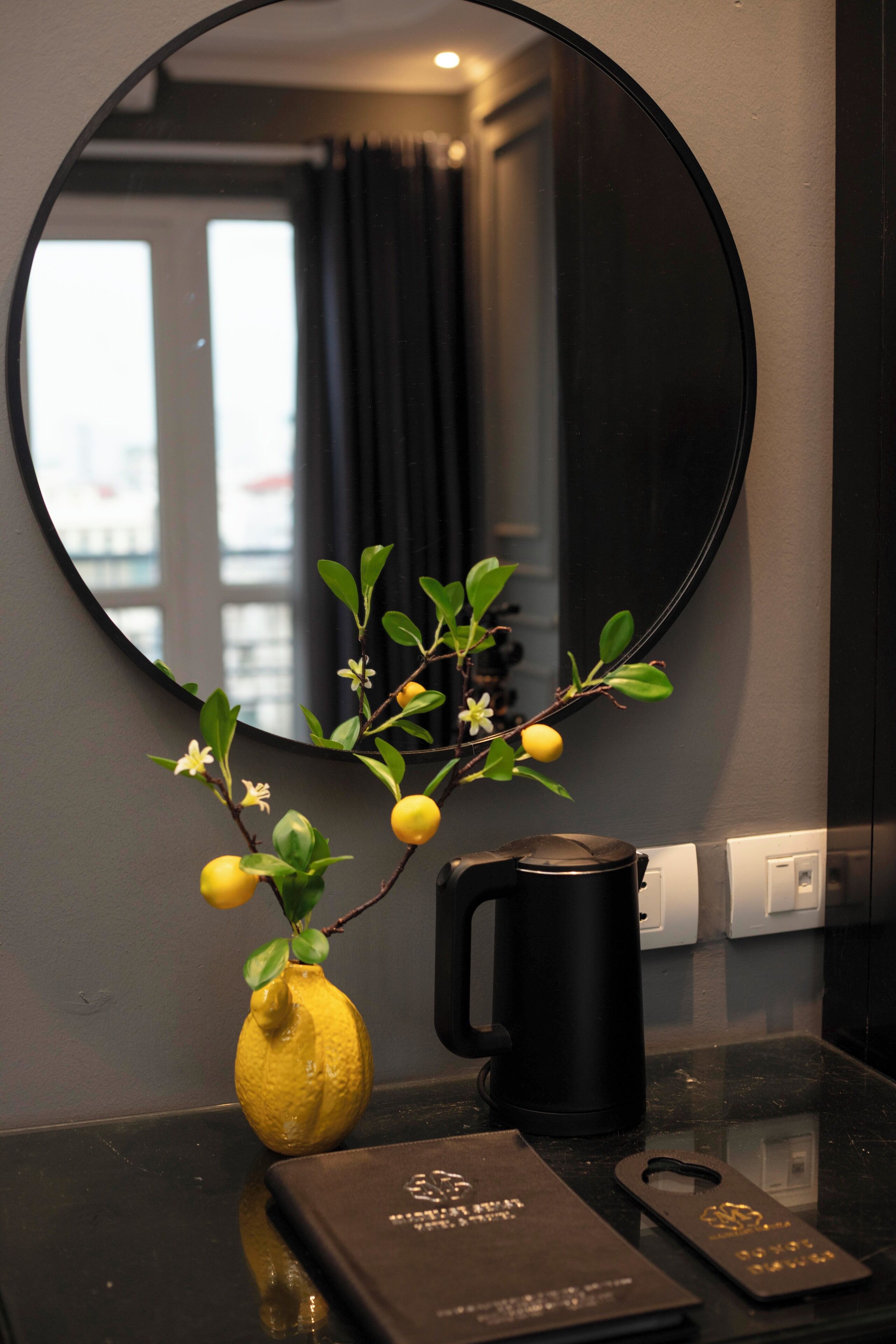 A bathroom with a round mirror, a yellow vase with a branch of flowers, a black mug, and a book on the counter.