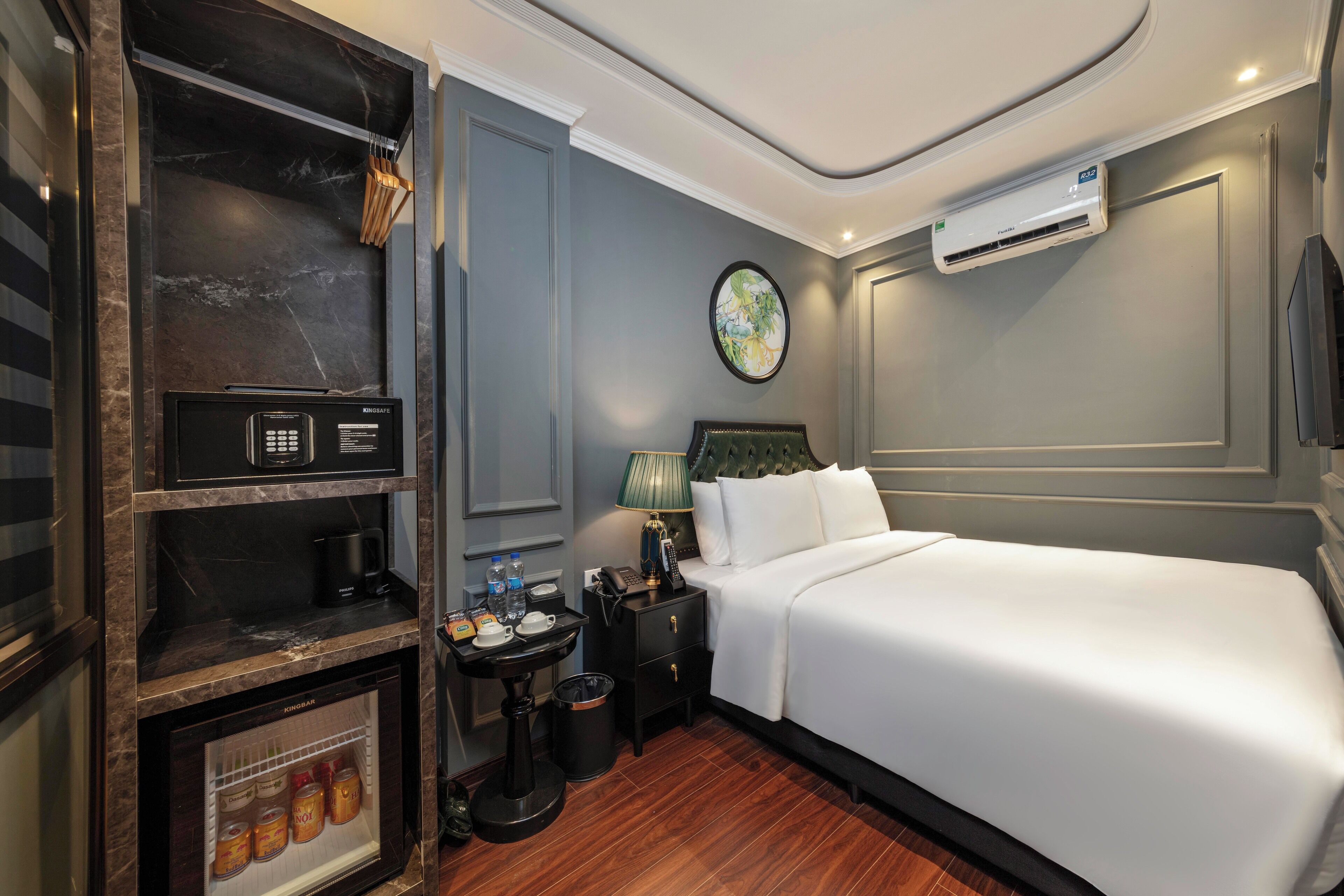 A hotel room with a bed, a minibar, a small table with a lamp, a trash bin, and a wall-mounted air conditioner.