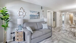 Ein Wohnzimmer mit grauem Sofa, einer Pflanze, einer Wandhalterung und einem Gemälde zum Thema Seefahrt.