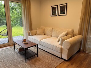 A living room with a beige sofa, a small table, and a view of a green garden through a glass door. - Nature Views & Sauna in the Eifel (Unknown)