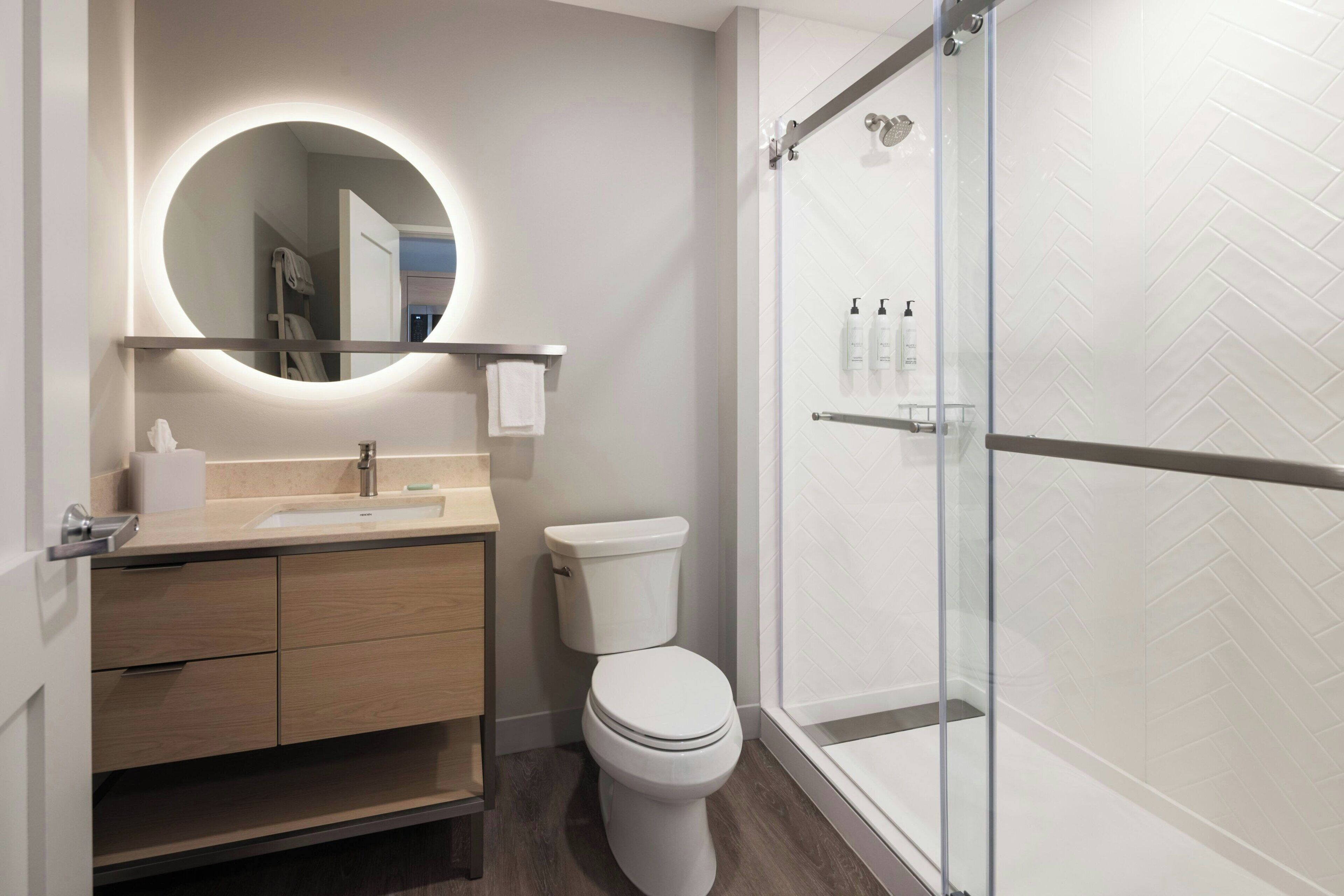 A modern bathroom with a round mirror, a toilet, a sink, and a glass-enclosed shower.