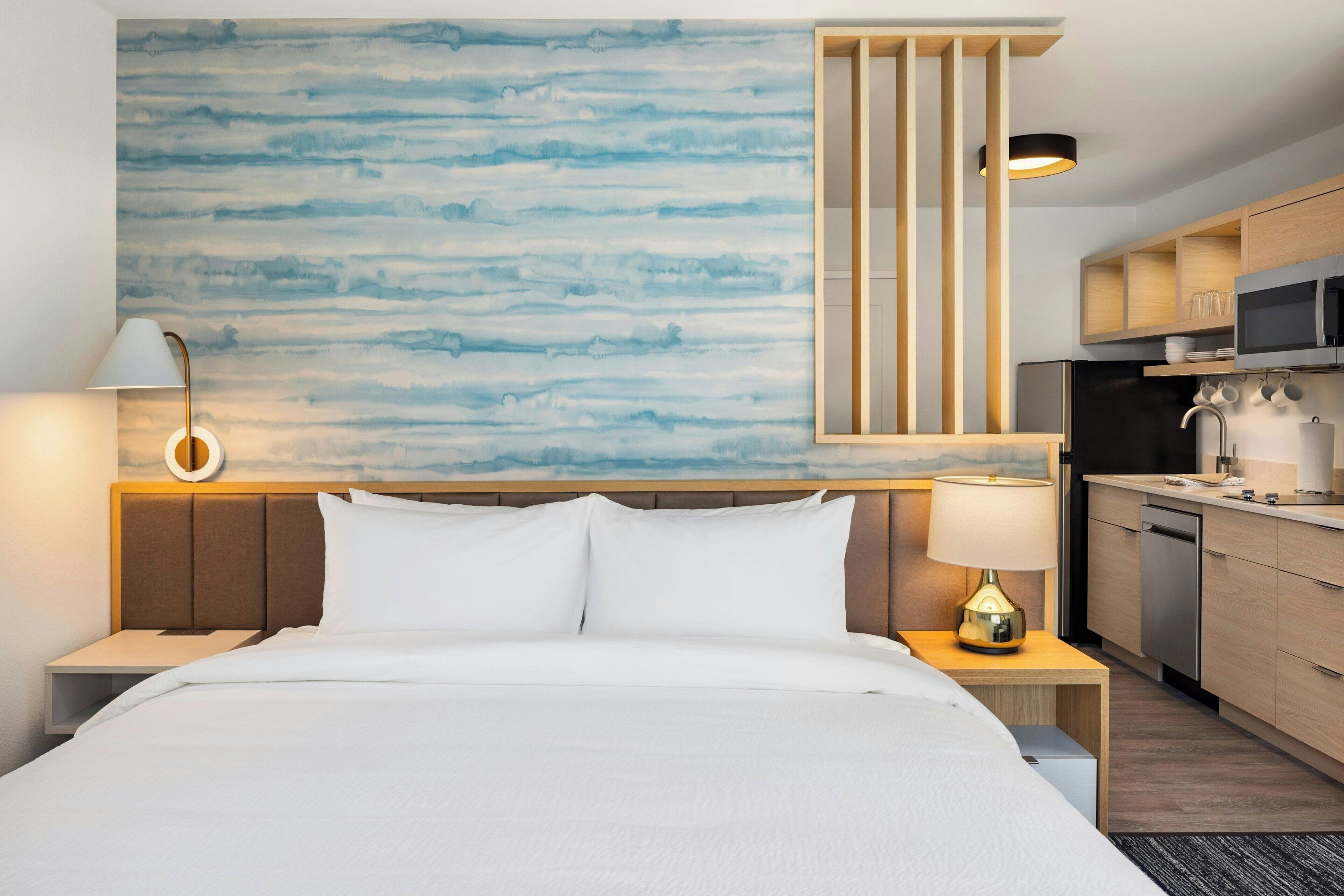 A hotel room with a bed, bedside tables, a wall-mounted lamp, a kitchen area with a microwave, and a dishwasher.