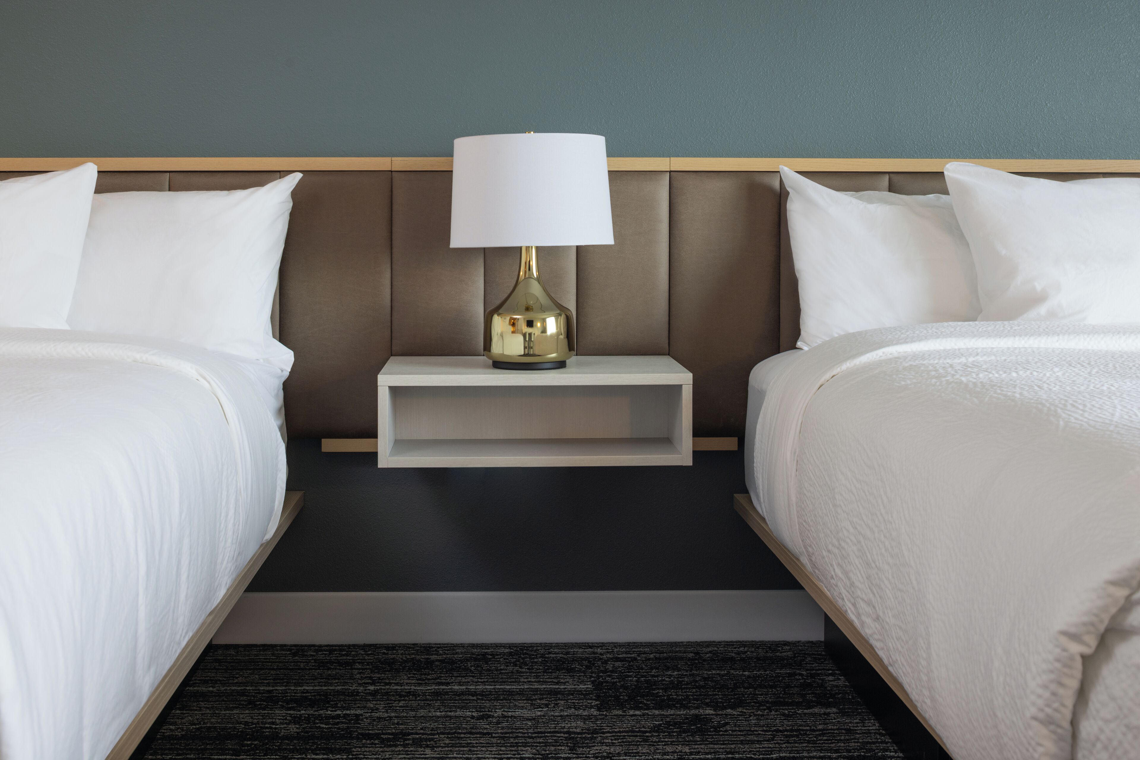 Una habitación de hotel con dos camas, una mesita de noche con una lámpara y una alfombra oscura.