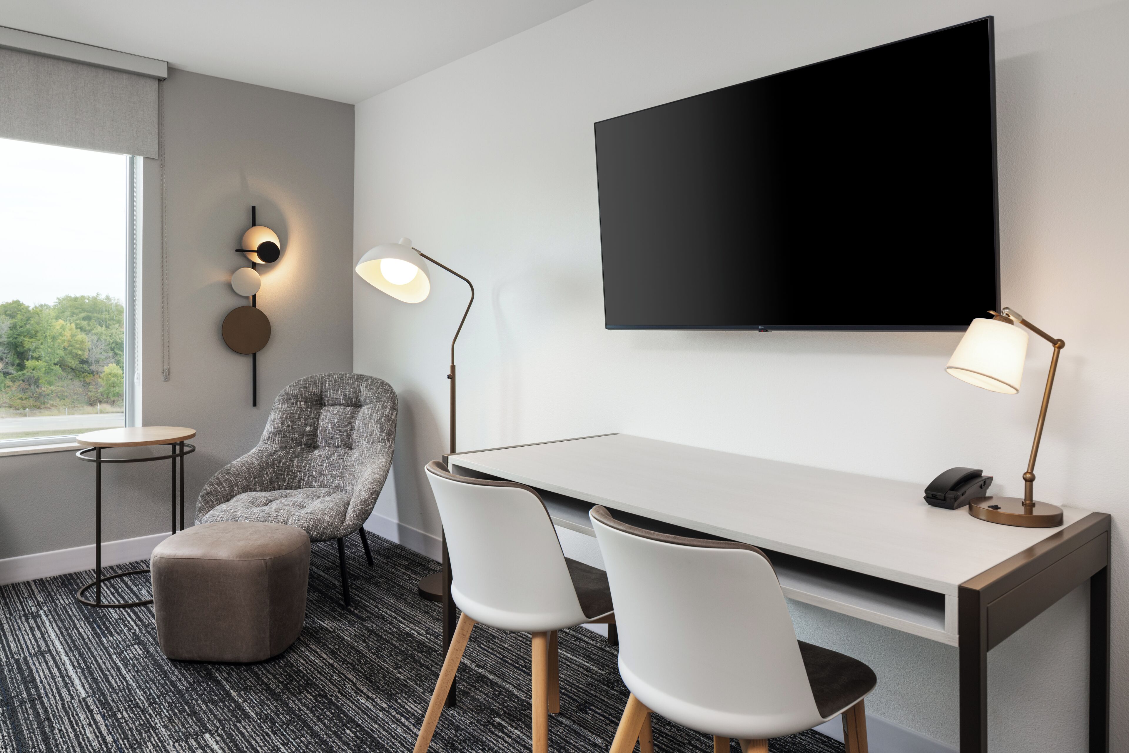 A modern hotel room with a large flat-screen TV mounted on the wall, a desk with two chairs, a grey armchair, a small round table, and a lamp.