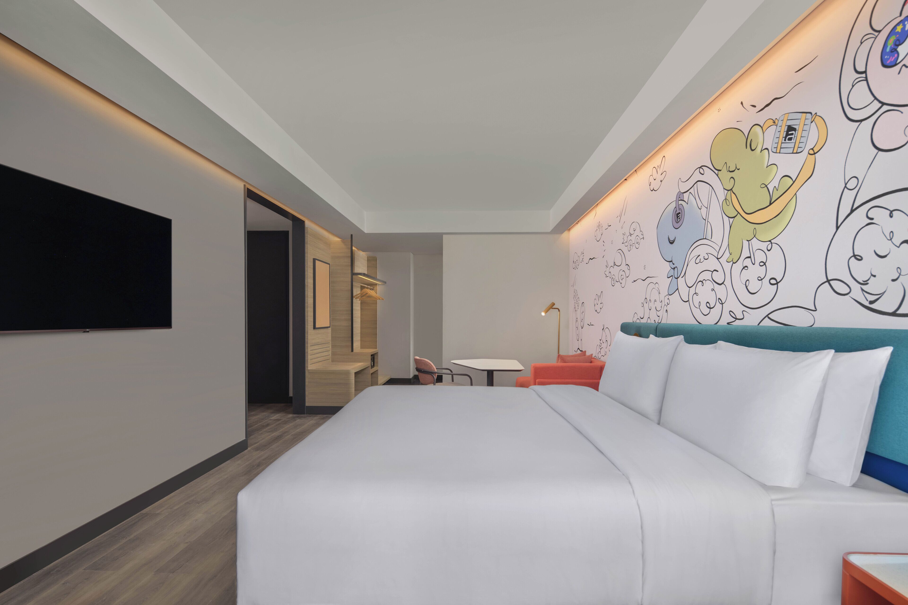 Una habitación de hotel moderna con una cama grande, un televisor de pantalla plana montado en la pared, una mesita pequeña y un mural colorido.