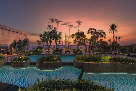 A rooftop pool with palm trees, lounge chairs, and a cityscape at sunset.. Aloft by Marriott Surabaya Pakuwon City