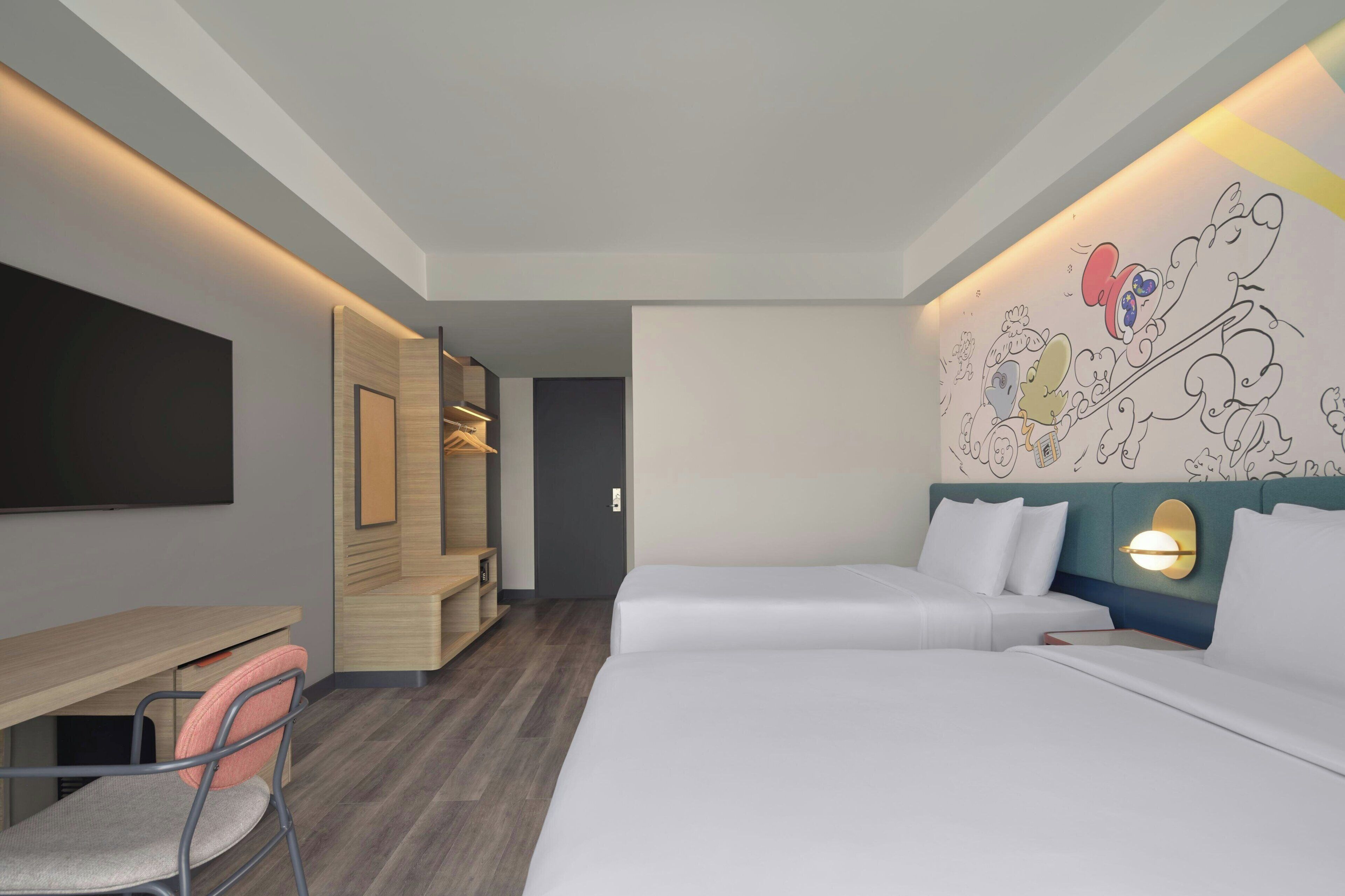 A modern hotel room with a desk, two beds, a chair, and a wall with cartoon characters.